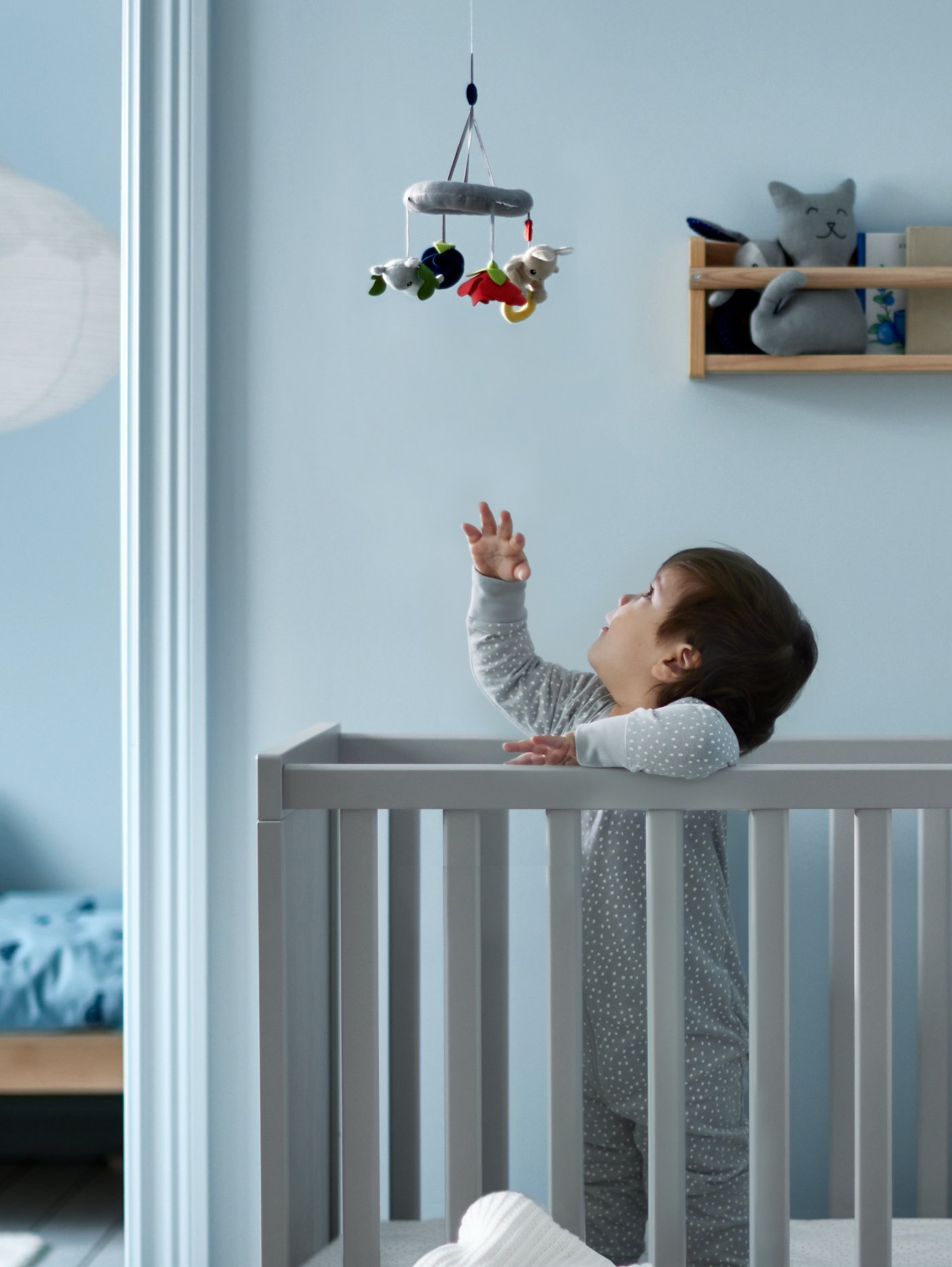 Un niño pequeño dentro de una cuna SUNDVIK intentando alcanzar un móvil que está sobre él colgando del techo.