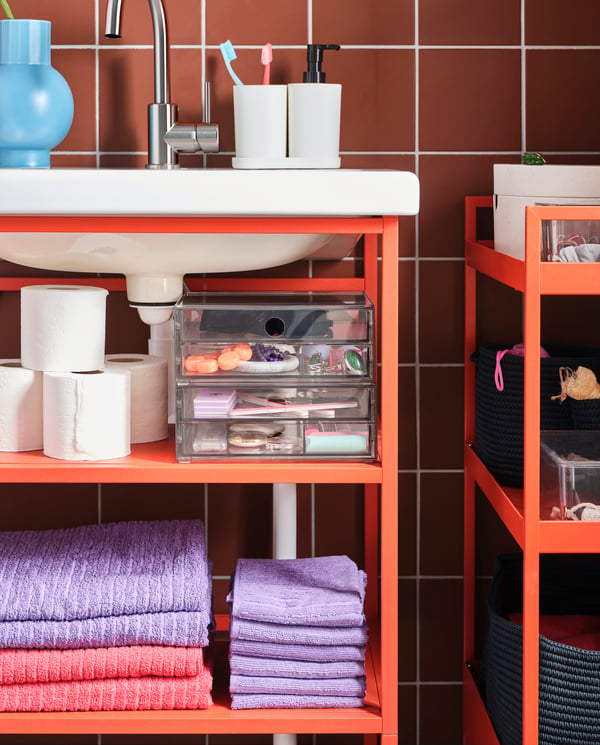 Vista de cerca de un mueble de baño ENHET abierto, de color rojo, y con el lavamanos empotrado en la parte superior. En el mueble se ven toallas dobladas, papel higiénico y cajas organizadoras transparentes.