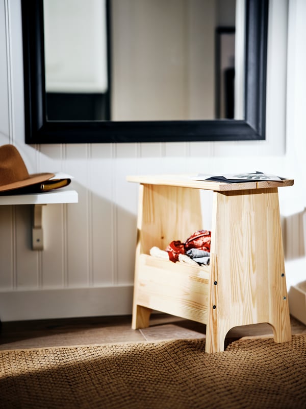 A pine PERJOHAN stool on a brown rug in a sunny space beside a low shelf and in front of a black TOFTBYN mirror.