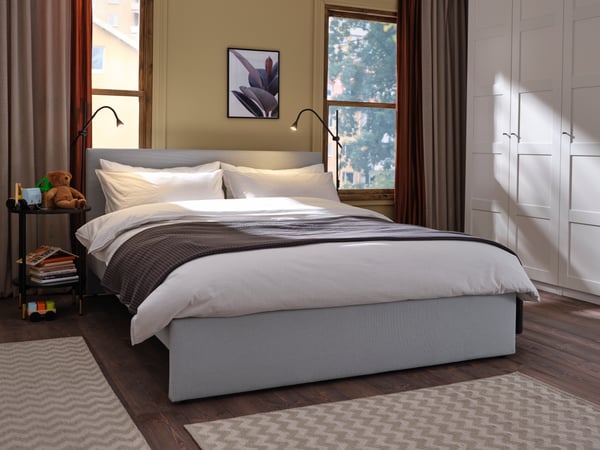 Cama tapizada GLADSTAD color gris, situada entre 2 ventanas con un plumón blanco y un cubrecama color gris.