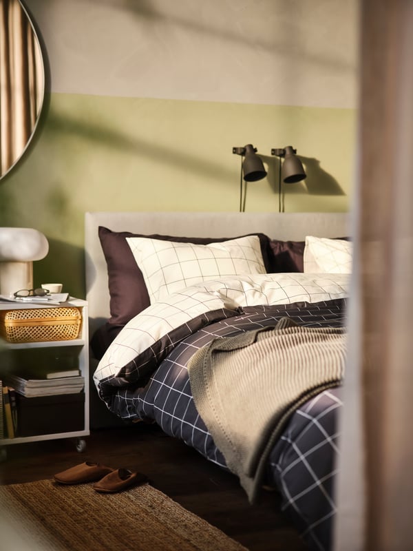 Nachtkastje met lamp, naast een lichtgrijs gestoffeerd GLADSTAD bed met daarop een wit-zwartgeruit VITKLÖVER dekbedovertrek.