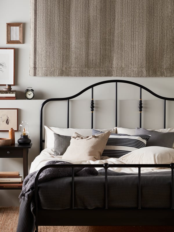 A black/Luröy SAGSTUA bed in a light room, covered in a mix of bed textiles and cushions in white, grey, black and beige.