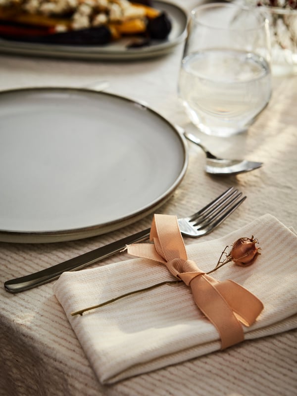 A table with a glass and a plate, spoon and fork beside a SILVERARV napkin with a ribbon holding a small festive bloom.