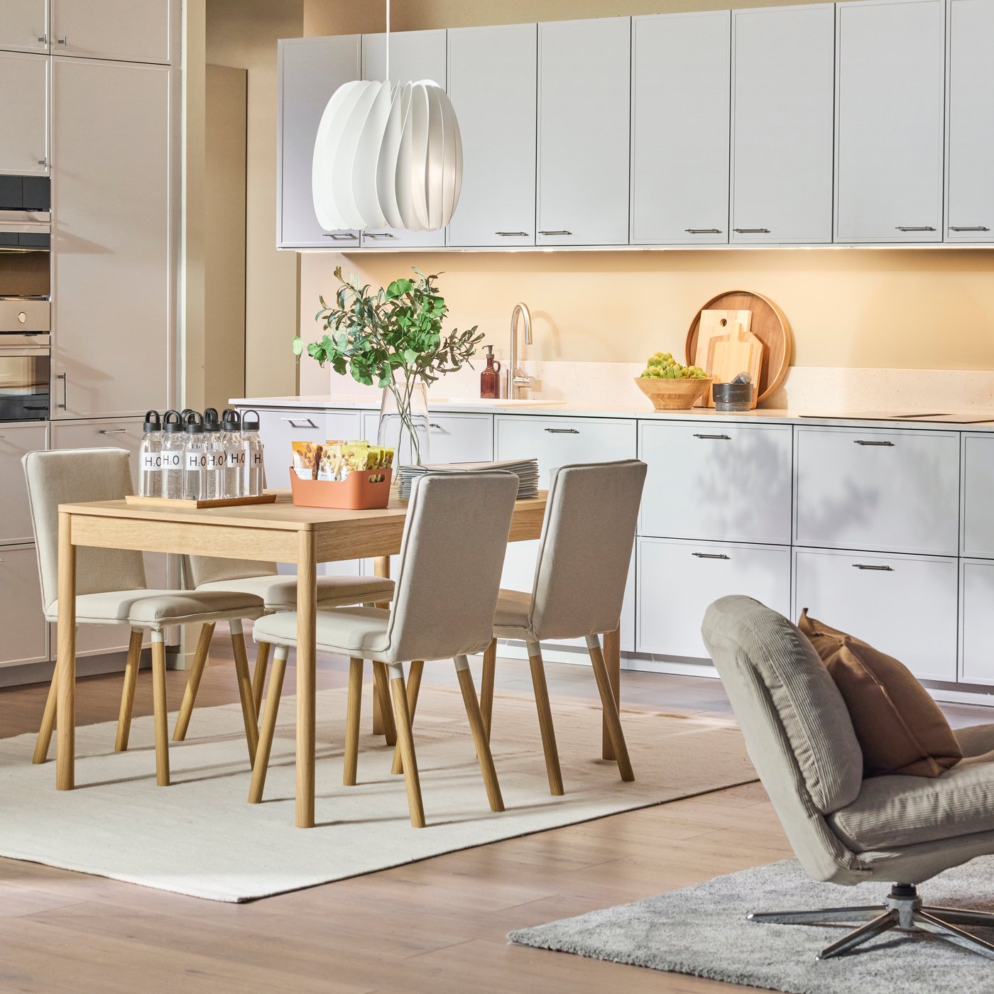 Ljus och modern köksinredning med vita skåp, matbord och stolar, taklampa, växter och prydligt ordnade föremål.