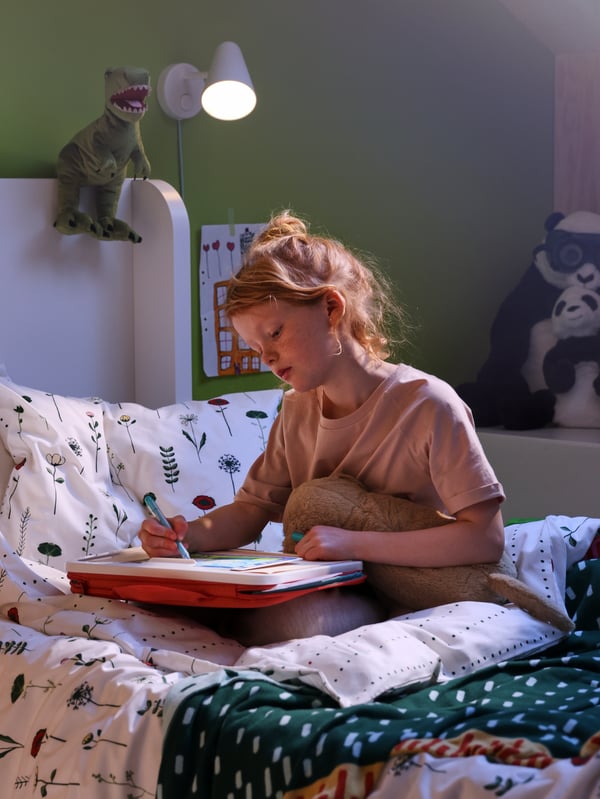 A child sits on her bed and draws with a MÅLA portable drawing case. A FUBBLA lamp and a JÄTTELIK soft toy are behind her.