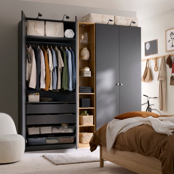 Un dormitorio con un armario PAX abierto, con puertas FORSAND en gris oscuro, ropa colgada y estantes en el interior.
