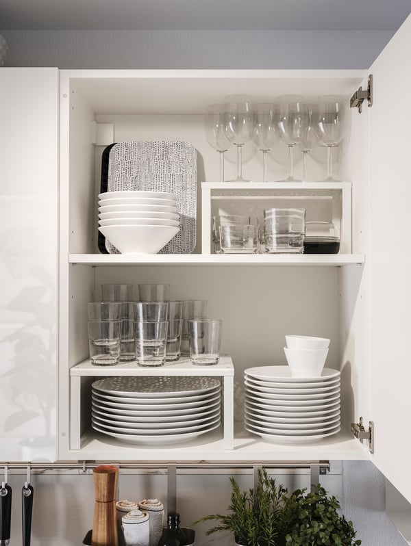 An opened KNOXHULT cabinet showing drinking glasses, wine glasses, plates, cups, bowls and two black and white trays.