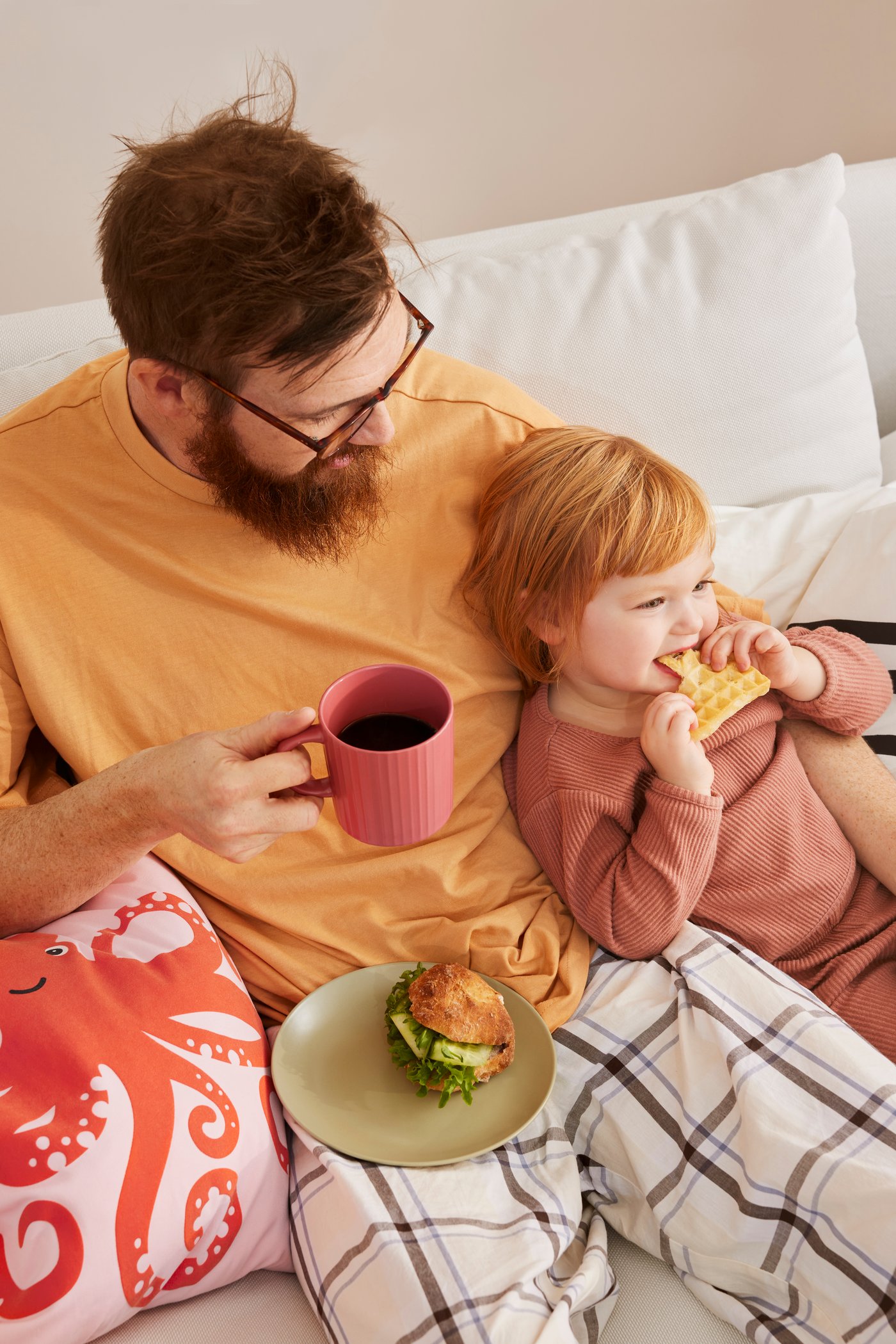 En pappa med sitt barn fikar i sängen.