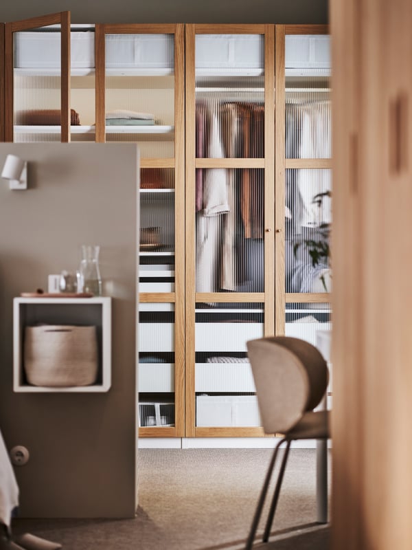 A white/oak veneer glass PAX/TONSTAD wardrobe combination filled with clothing with a dark beige KRYLBO chair in front.