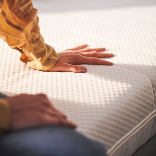 A person with a yellow shirt sitting on the corner of two white ÅKREHAMN foam mattresses. One hand is touching the surface.