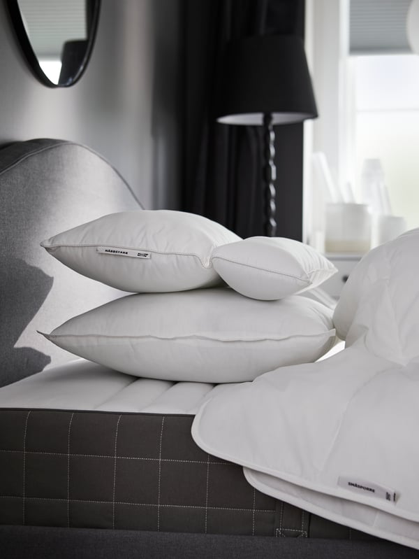 Un oreiller NÄBBSTAR ergonomique a été posé au sommet du lit, près d'un autre oreiller et d'une couette SMÅSPORRE, et un miroir rond est fixé au mur.