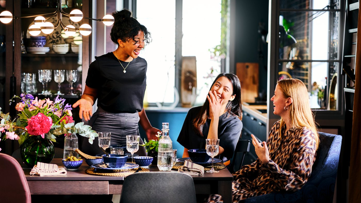 Three women are chatting and laughing around a dining table set with blue dinnerware, a sunny kitchen in the background.