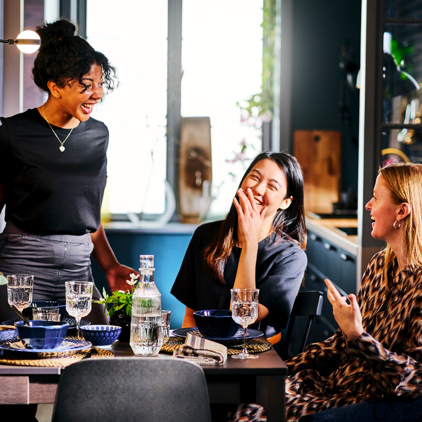 Three women are chatting and laughing around a dining table set with blue dinnerware, a sunny kitchen in the background.