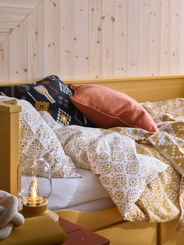 Žltá posteľ HEMNES pri nočnom stolíku s lampičkou TÄRNABY a s bielym vzorovaným povlečením ORMÄNGSFLY.
