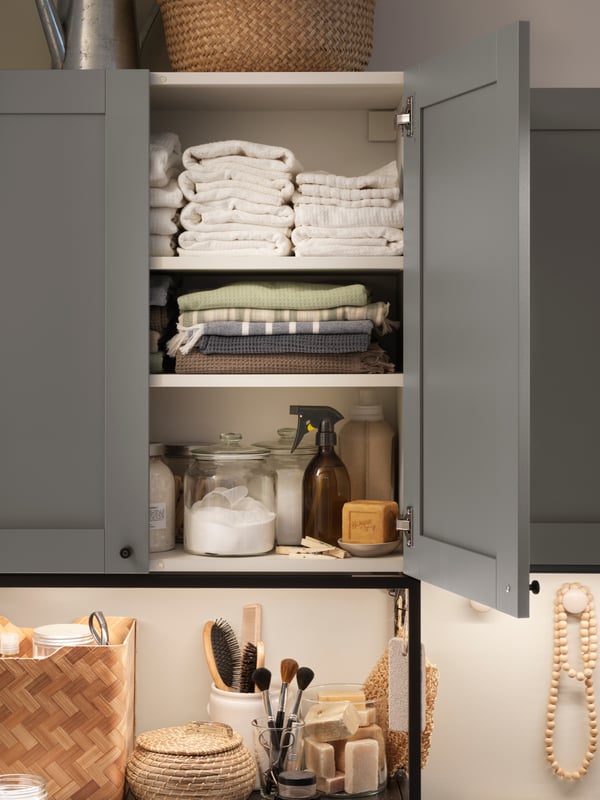 A grey ENHET bathroom cabinet, door open, inside are diverse glass jars and towels with bathroom items on the shelf below.