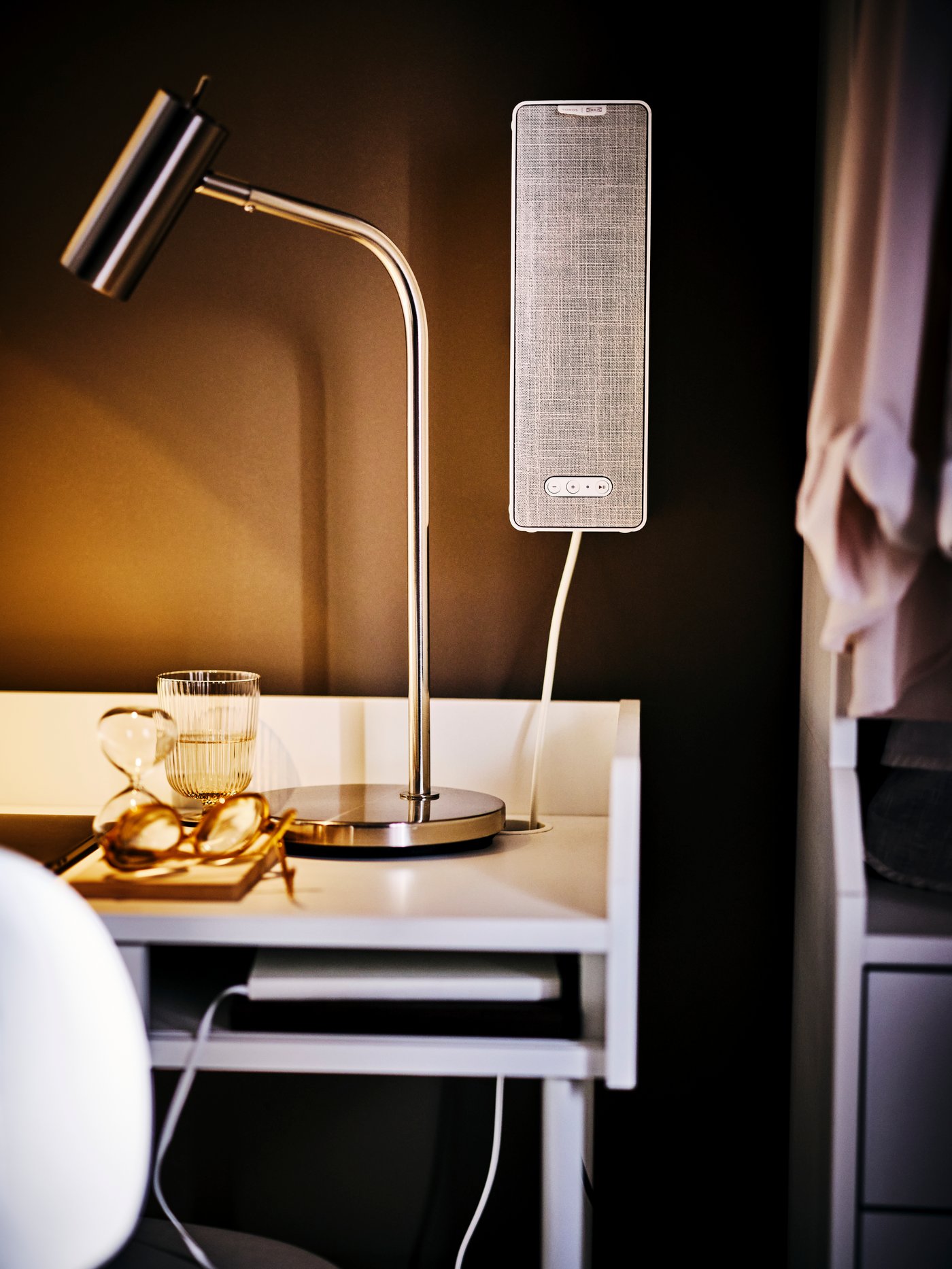 The right corner of a white HAUGA desk with a lit VIRRMO work lamp and a SYMFONISK WiFi bookshelf speaker on the wall.