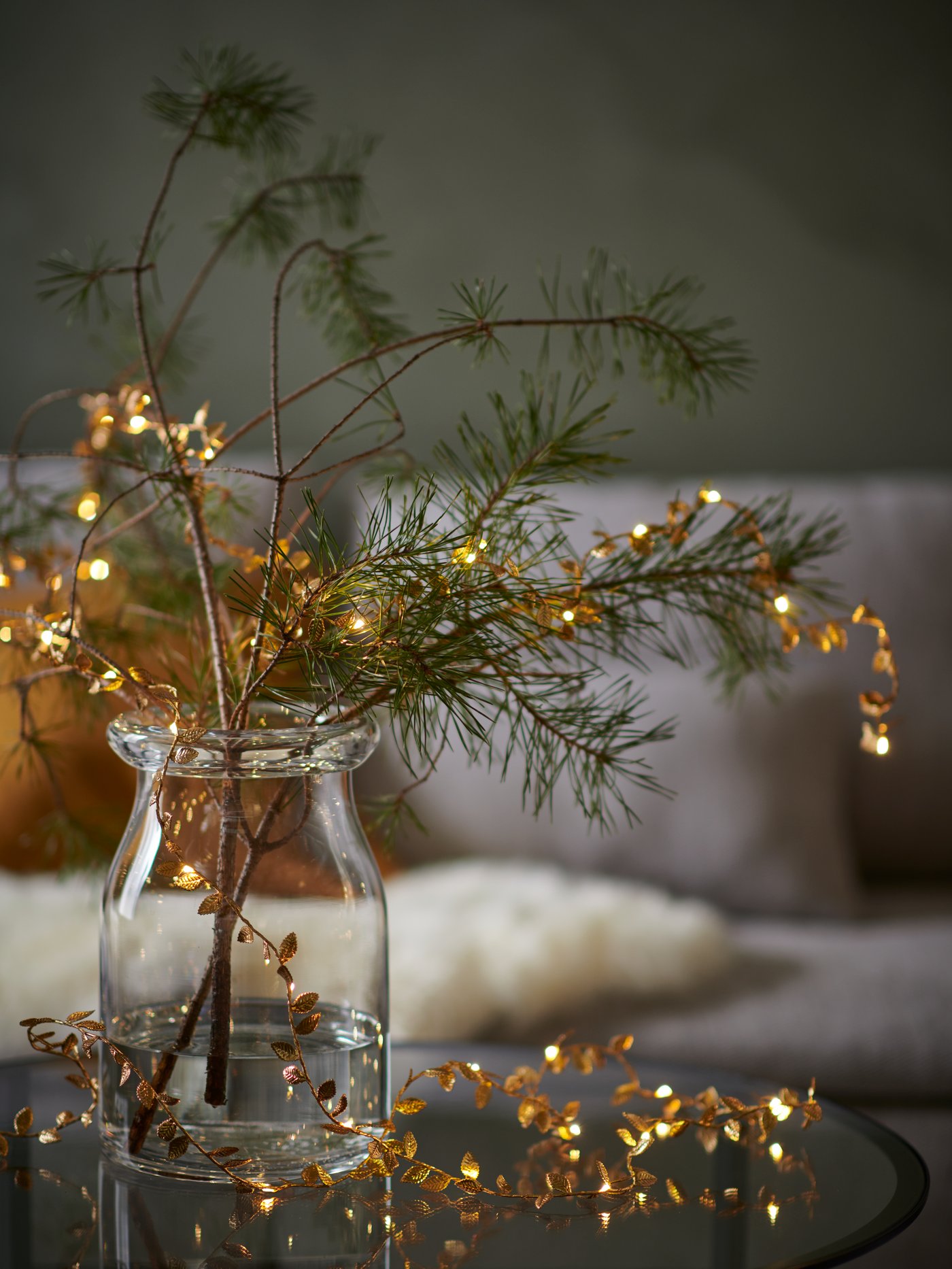 Pine branches decorated with golden STRÅLA LED lighting chains stand in a BEGÄRLIG clear vase on a small glass tabletop.