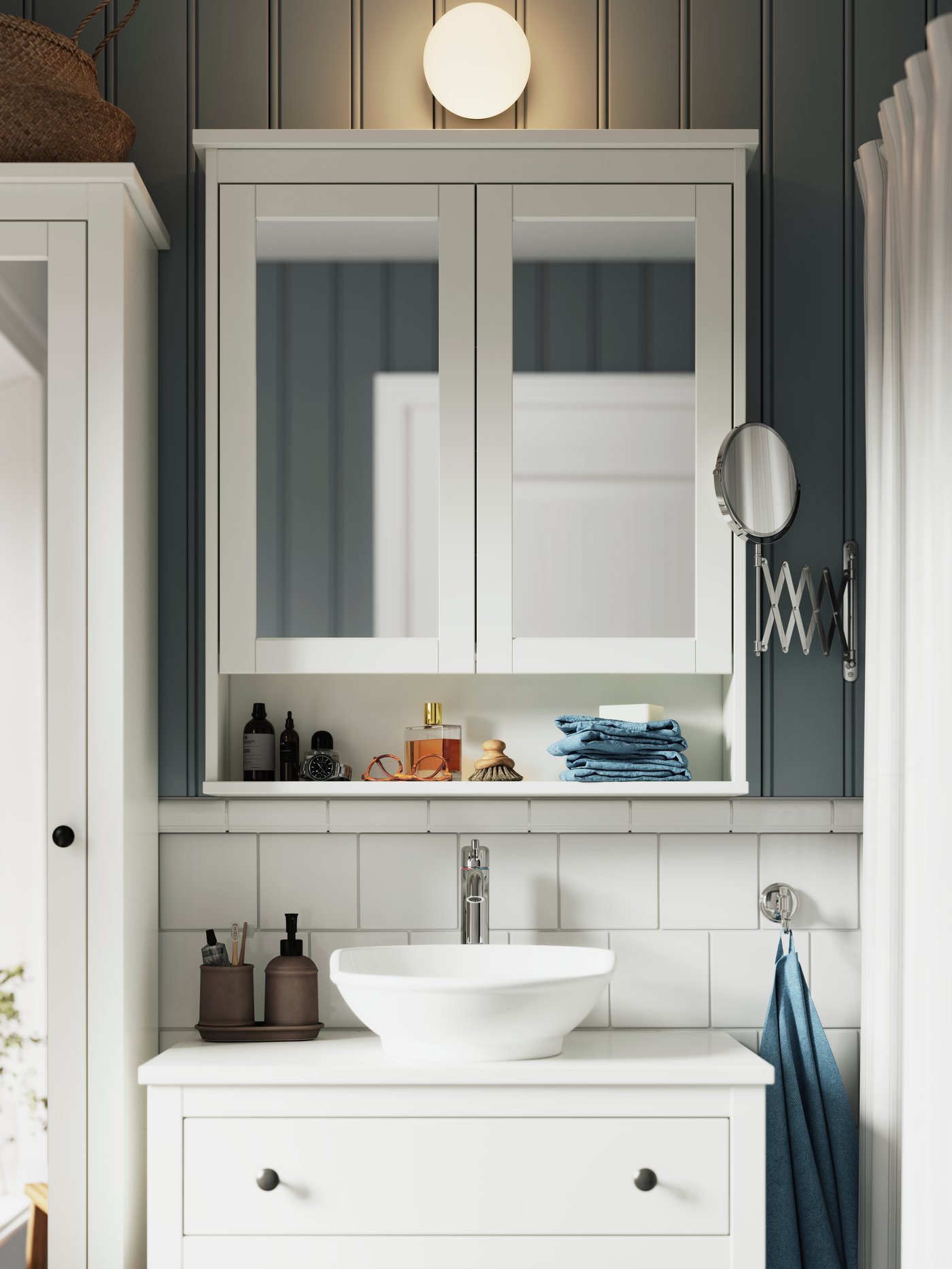 Nad bílou zrcadlovou skříňkou HEMNES a bílým umyvadlovým stolkem HEMNES je namontována nástěnná lampa FRIHULT z kovu a skla.