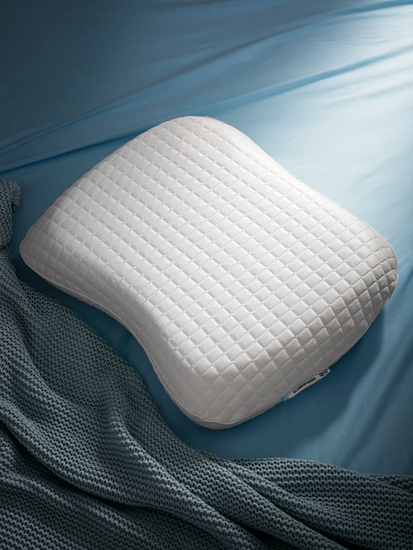 An uncovered KLUBBSPORRE pillow on a bed covered with blue bed linen and a grey-blue blanket.
