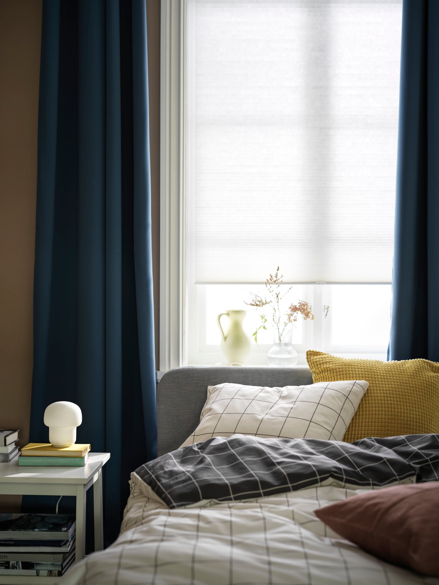 La cabecera de una cama junto a una ventana con un tratamiento de ventana compuesto por una cortina de velo HOPPVALS y cortinas de tela HILLEBORG.