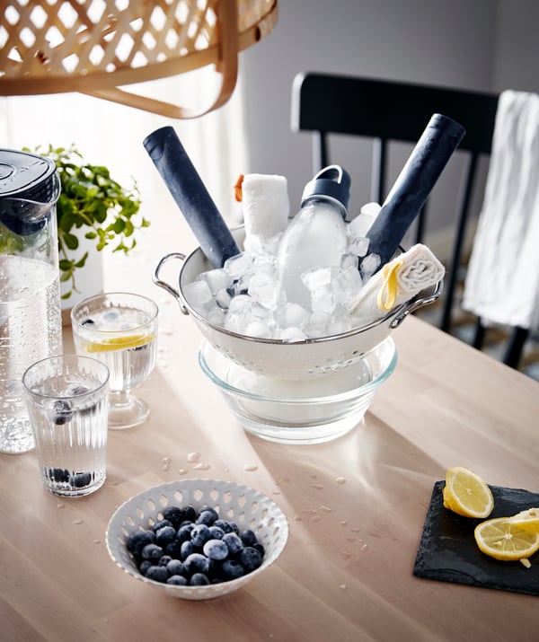Bilden visar ett bord med olika föremål på. Det finns en glasskål fylld med is och flera flaskor, en liten skål med blåbär, två glas vatten med citronskivor och en svart servett med två citronklyftor på. I bakgrunden syns en stol och en del av en annan möbel.