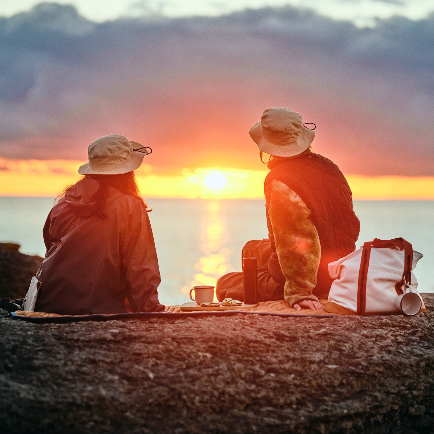 Två personer med SOLUPPGÅNG-hattar sitter på en SOLUPPGÅNG-picknickfilt och tittar på solnedgången vid havet.