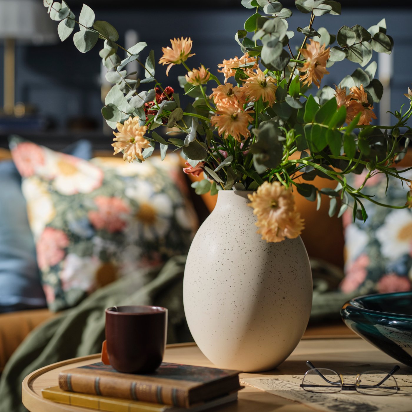 A BLODBJÖRK vase in beige stands with flowers on a coffee table in front of a sofa with cushions in ARENBOK cushion covers.