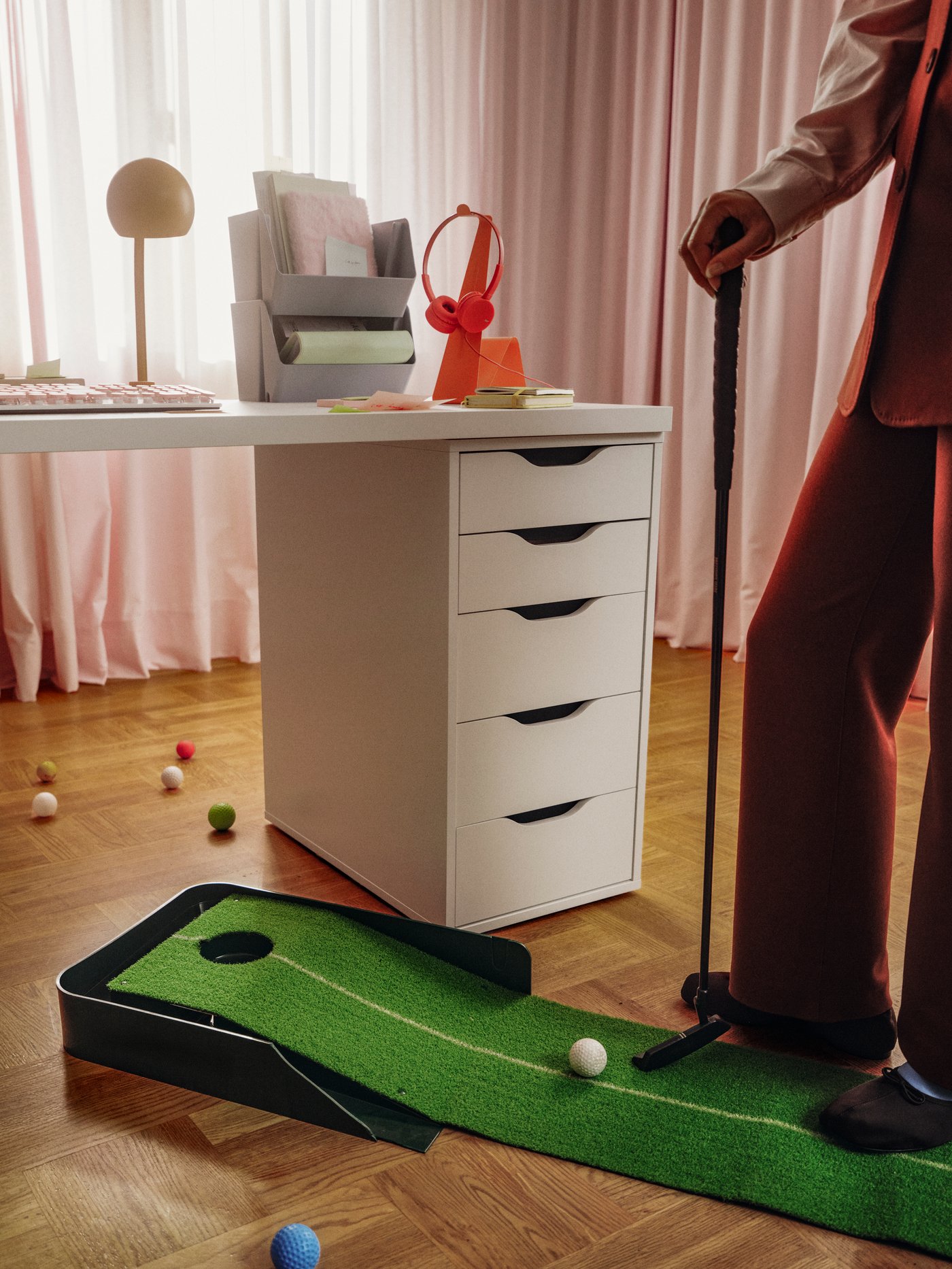 A white ALEX drawer unit with sleek, cut-out handles supports a white desk. Next to it, a person practices golf putts.