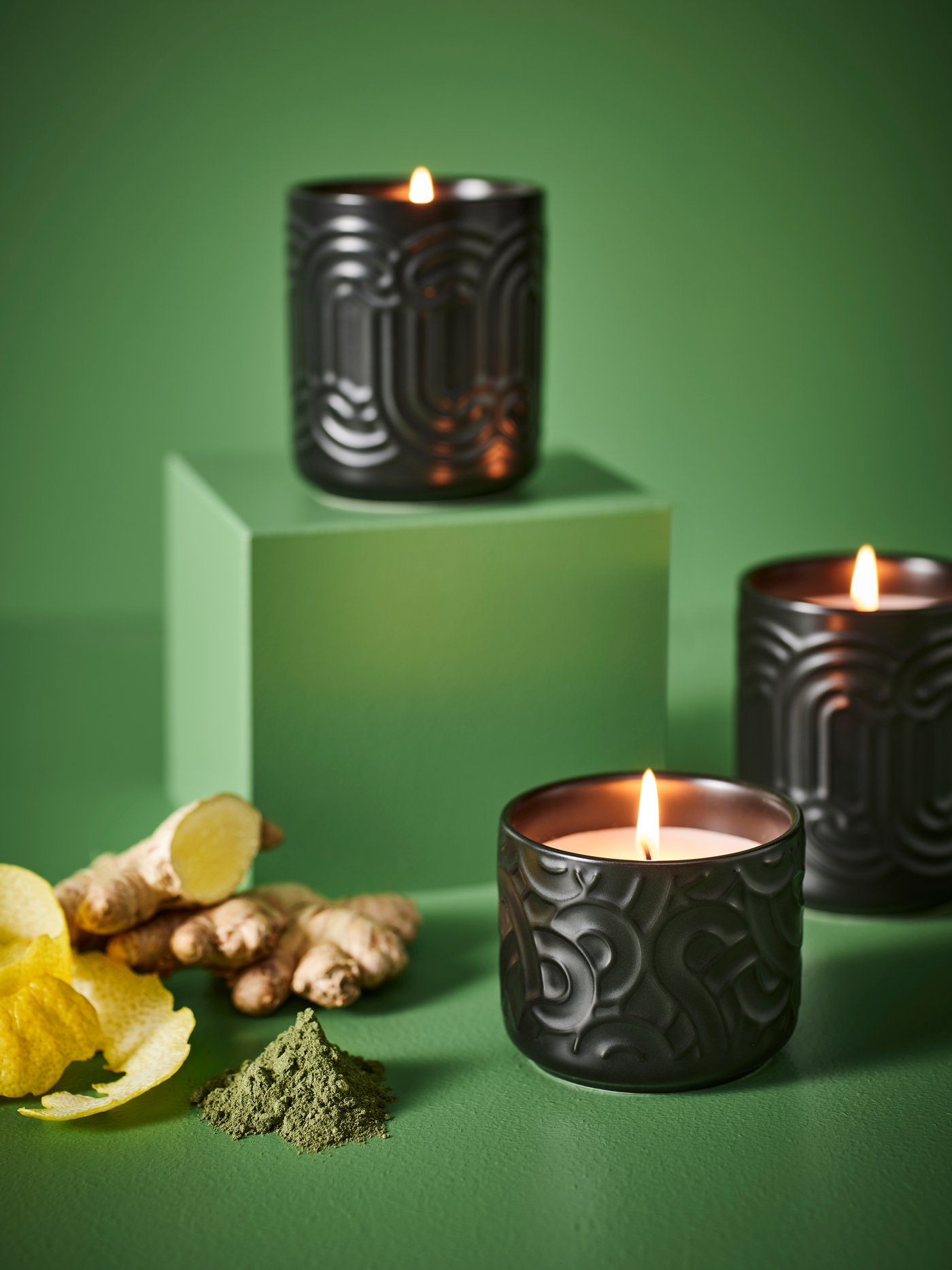 Three SÖTRÖNN scented candles in a green space. One is on a pedestal. There are ginger and lemon peel next to the pedestal.