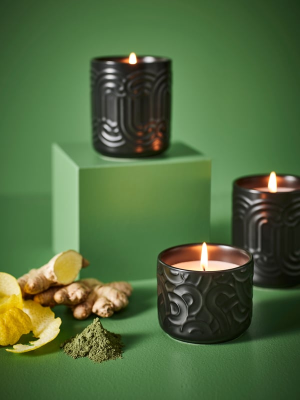 Trois bougies parfumées SÖTRÖNN dans une pièce verte. L’une d’entre elles est posée sur un socle. Ce dernier est entouré d’écorces de gingembre et de citron.