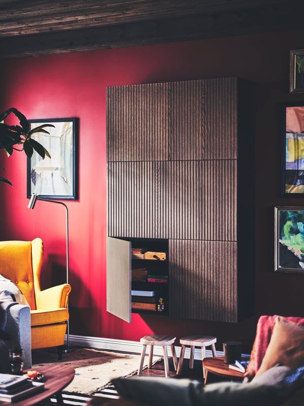 Una sala de estar con un mueble de almacenamiento con puertas BJÖRKÖVIKEN de color café, con dos taburetes debajo y un sillón amarillo STRANDMON.