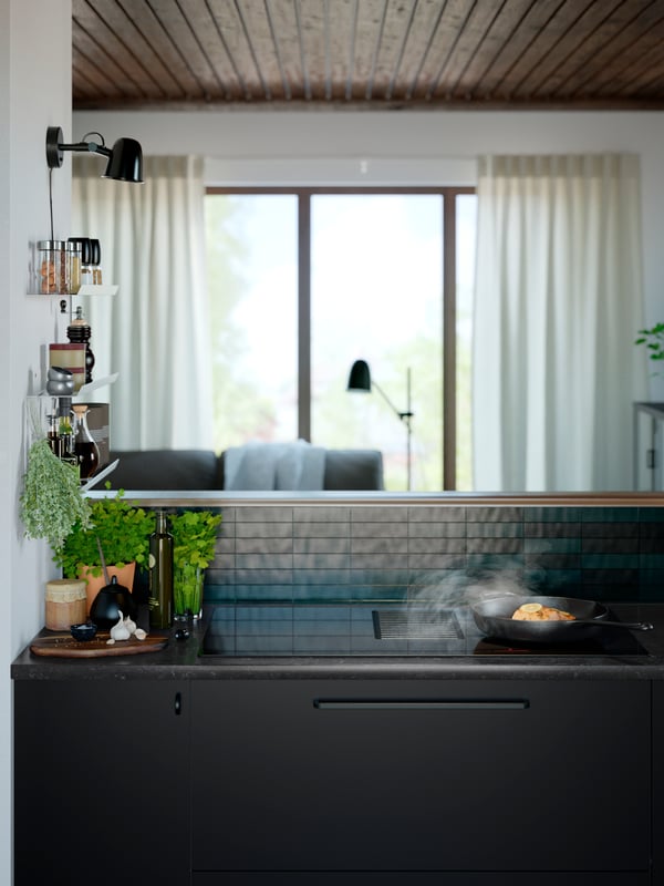Food is being prepared in a cast iron frying pan on a FÖRNEBO induction hob next to a pot of herbs and bottle of oil.