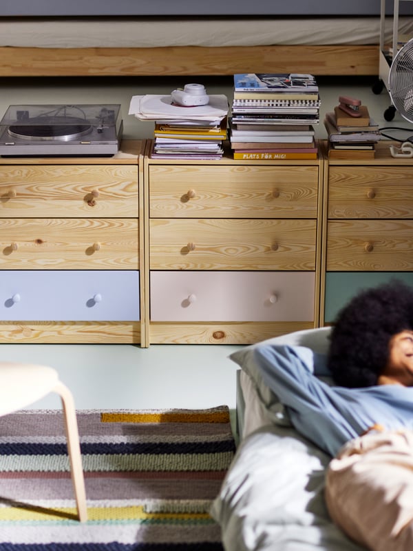 Three pine RAST chests of drawers with the bottom drawers painted in different colors, beside a woman lying on a bed.