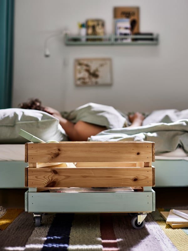 A partly painted KNAGGLIG pine box on castors on a multicolor rug by a bed with green bed linen where a man is sleeping.