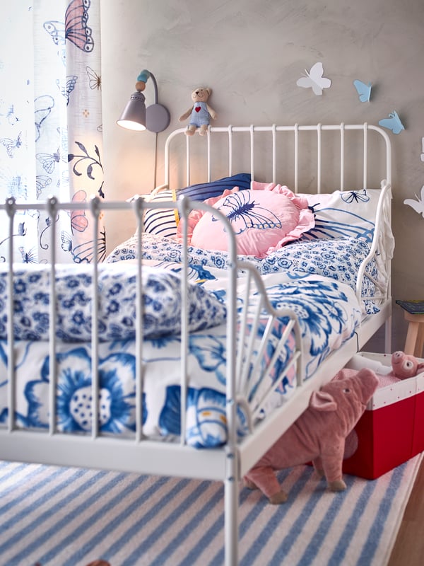 Un lit extensible MINNEN blanc garni de linge de lit bleu et blanc. Au sol, un tapis rayé et un cochon en peluche.