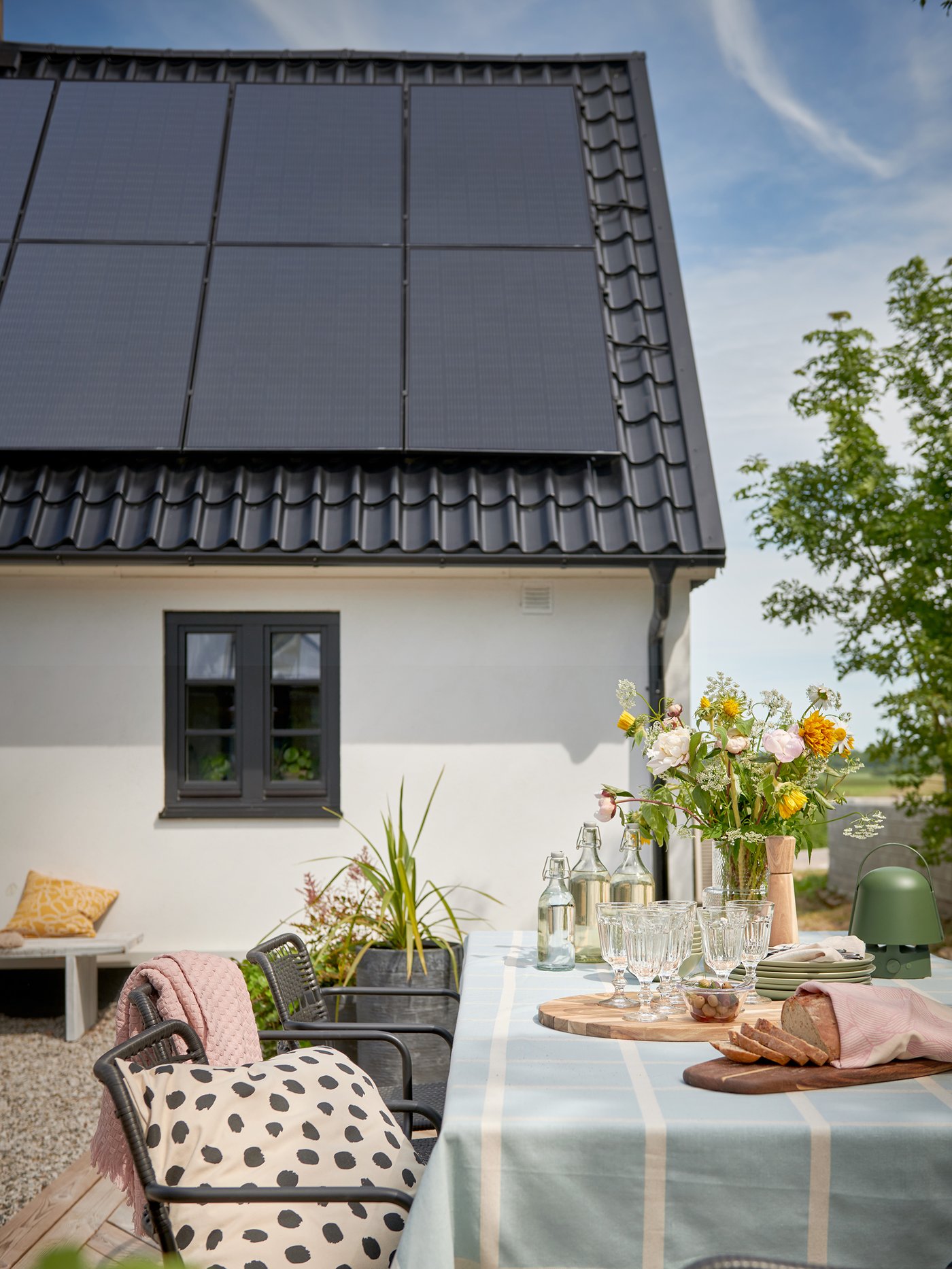 Uteplats vid ett ljust hus med solpaneler på taket. Ett dukat bord står i förgrunden med blommor, glas, vattenkaraffer och bröd. Runt bordet finns stolar med kuddar, och i bakgrunden syns ett fönster samt en bänk med kuddar. En grön trädgren sträcker sig in från höger under en klar himmel.