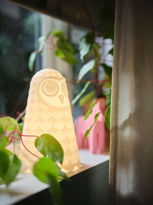 Uključena bela SOLBO LED stona lampa u obliku sove nalazi se na simsu pored prljavobele LENDA zavese s vezicom.