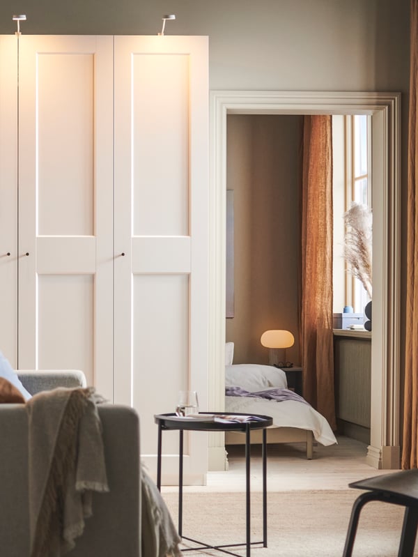 A PAX/GRIMO wardrobe with YTBERG cabinet lighting from an YTBERG/TRÅDFRI lighting kit stands beside a doorway into a bedroom.