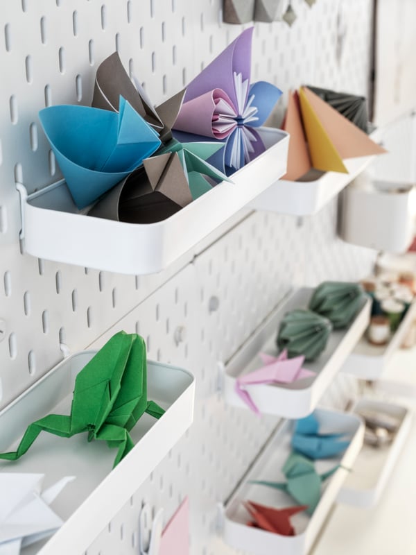 A patchwork of wall-mounted white SKÅDIS pegboards fitted with white SKÅDIS shelves, filled with origami objects.