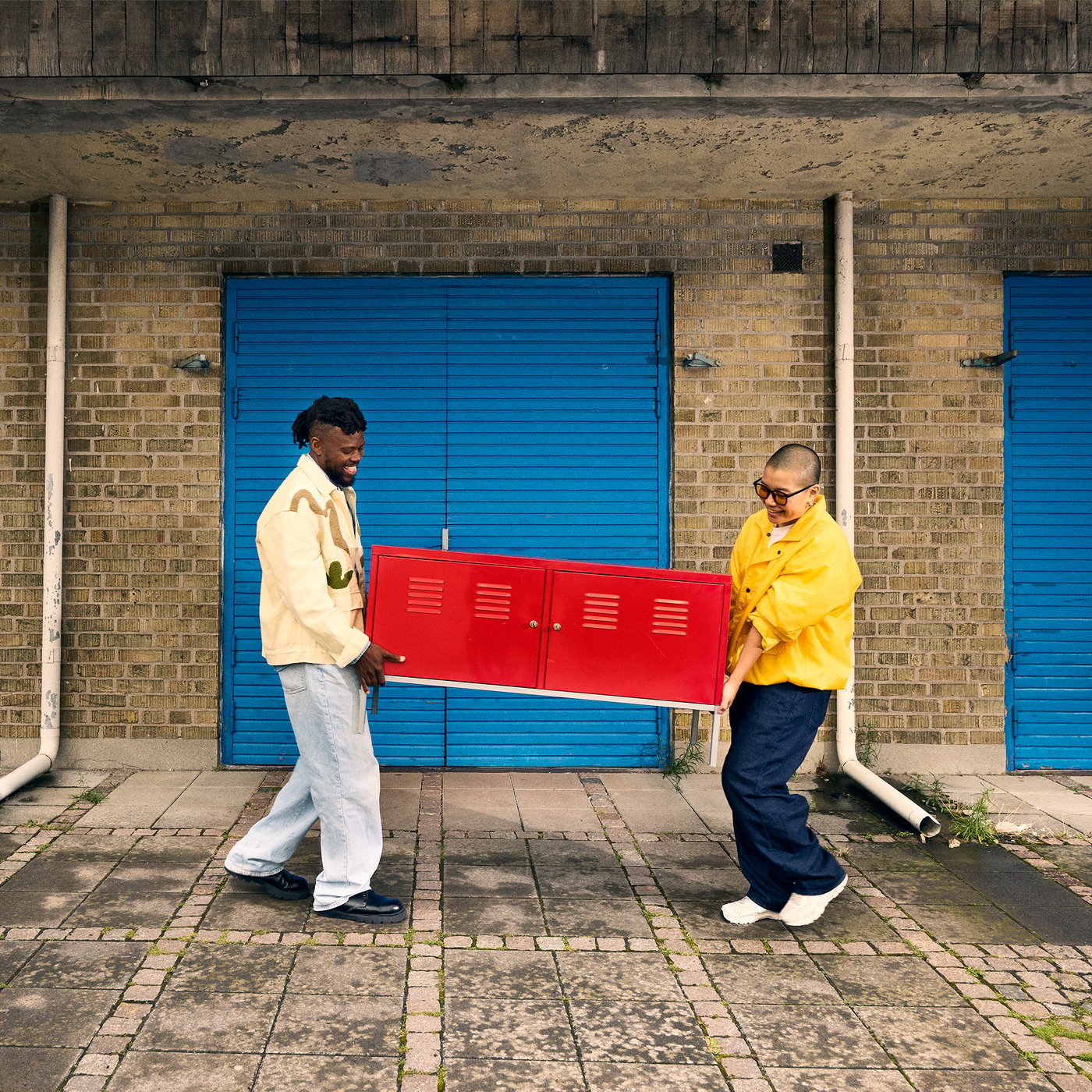 Two people are carrying a bright red locker outdoors in front of a brick building with blue doors.