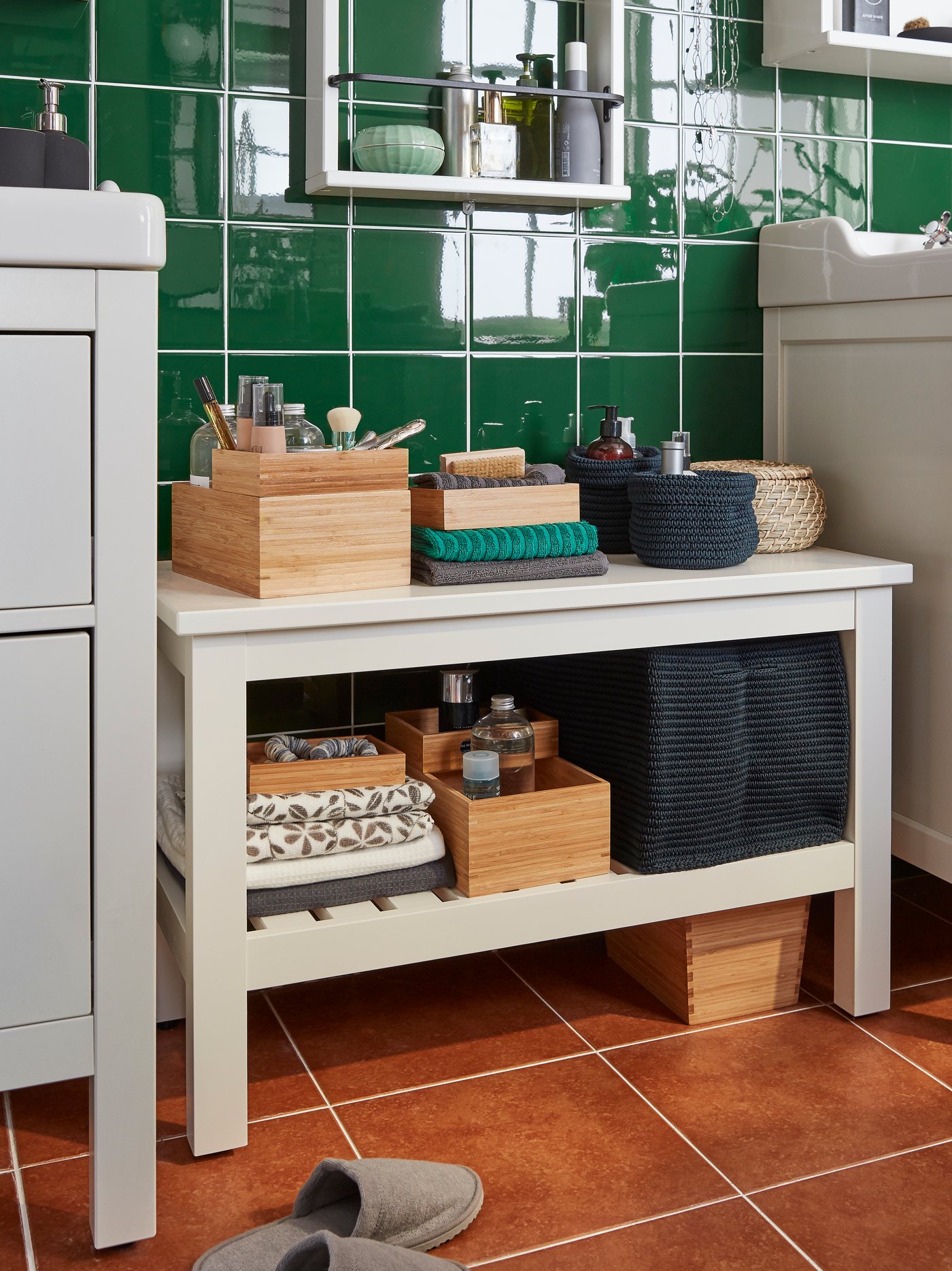 A HEMNES bench stands between two wash-basins in a green tiled bathroom. Boxes and towels are stored on the shelves.