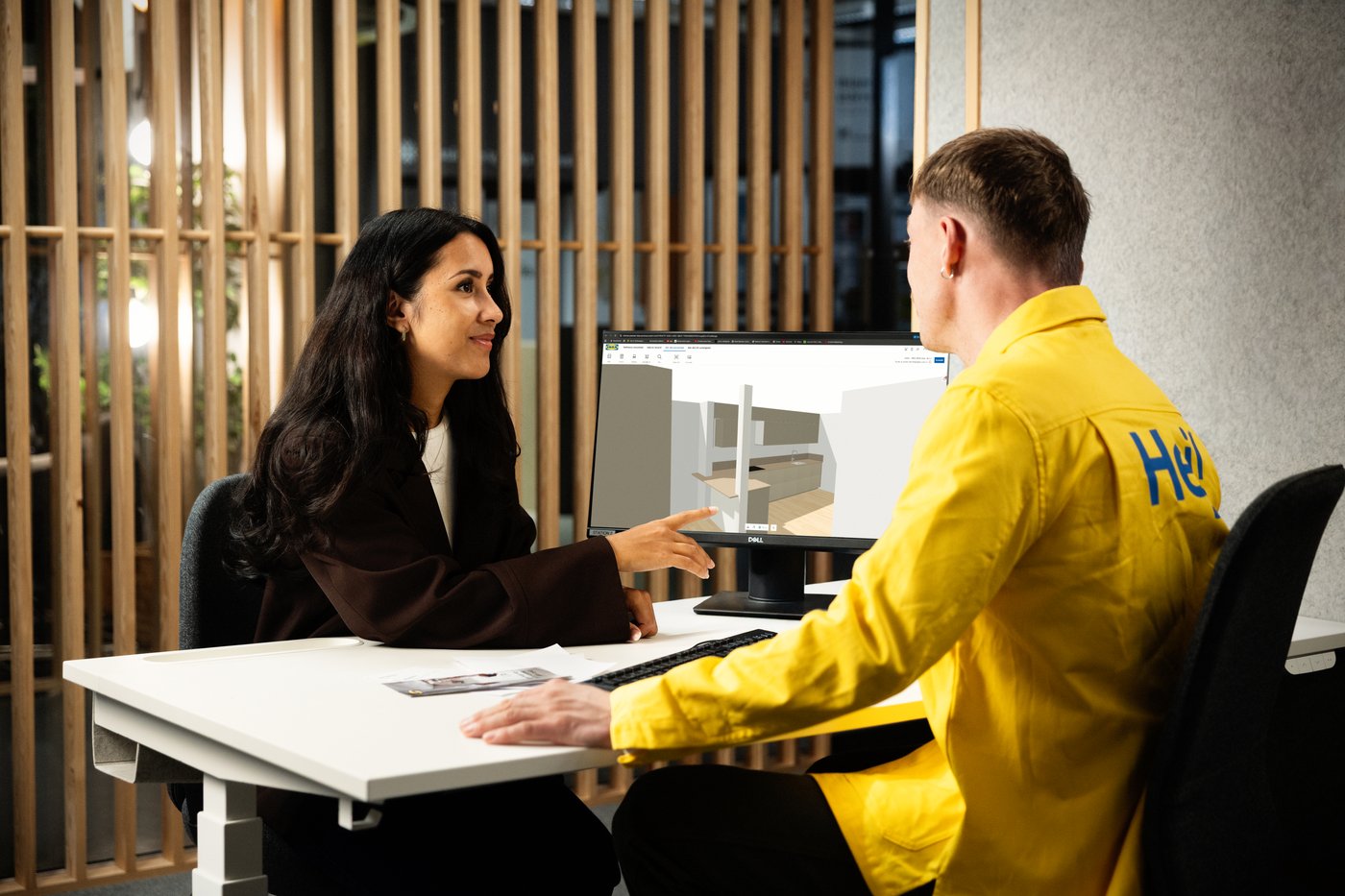 Two people are sitting at a desk discussing a digital kitchen plan on a computer screen.