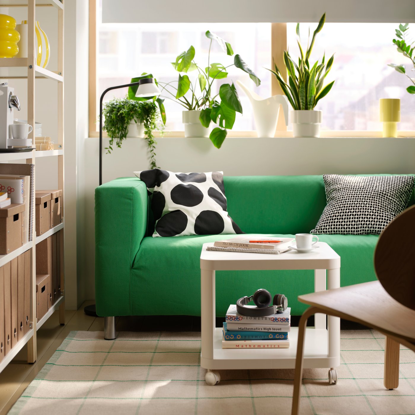 A Vissle green KLIPPAN two-seat sofa is in a minimally decorated student room rental, below potted plants on a window ledge.