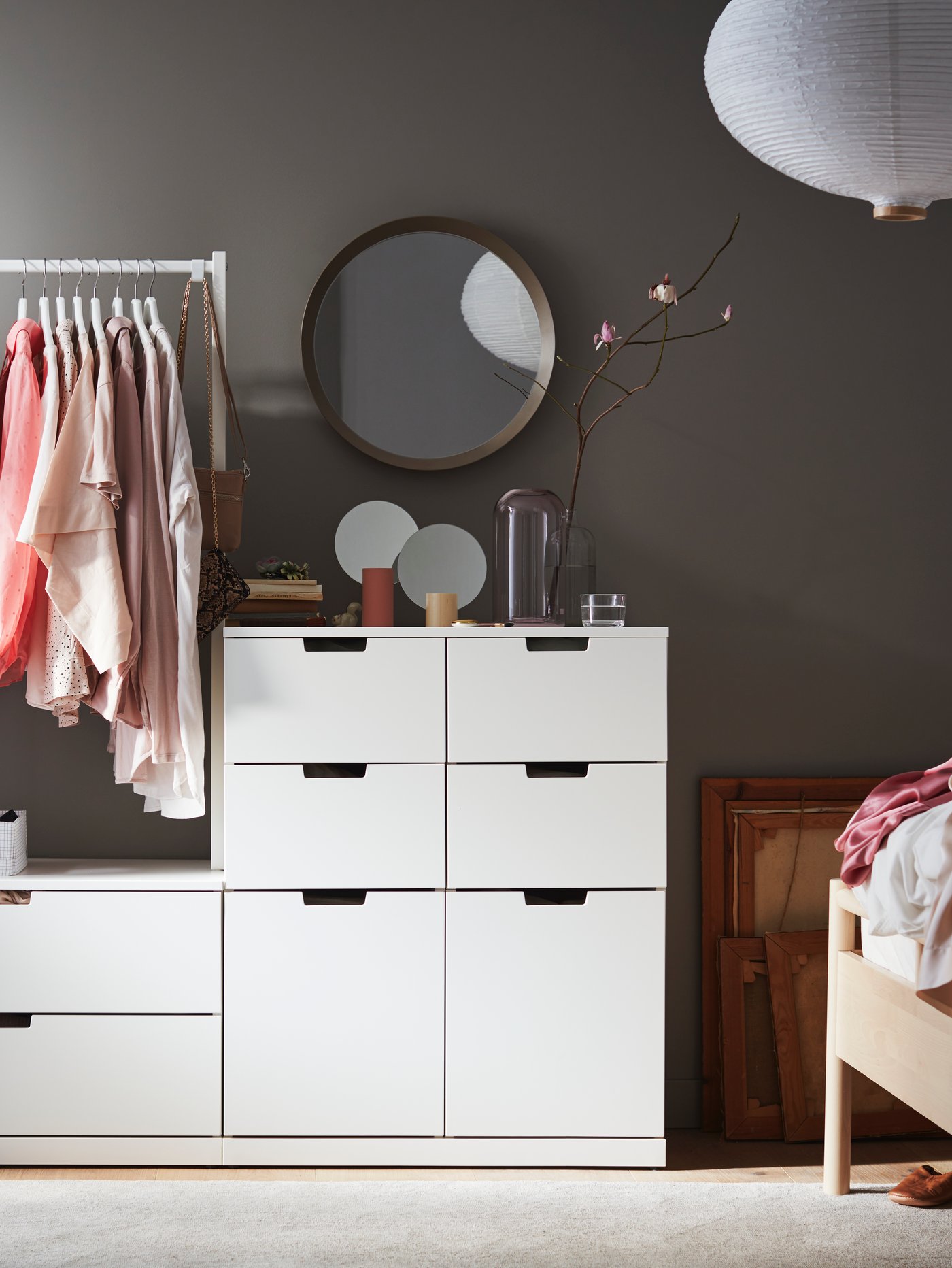 A NORDLI chest of 8 drawers with a clothes rail holding hanging clothes stands against a wall under a LANGESUND mirror.