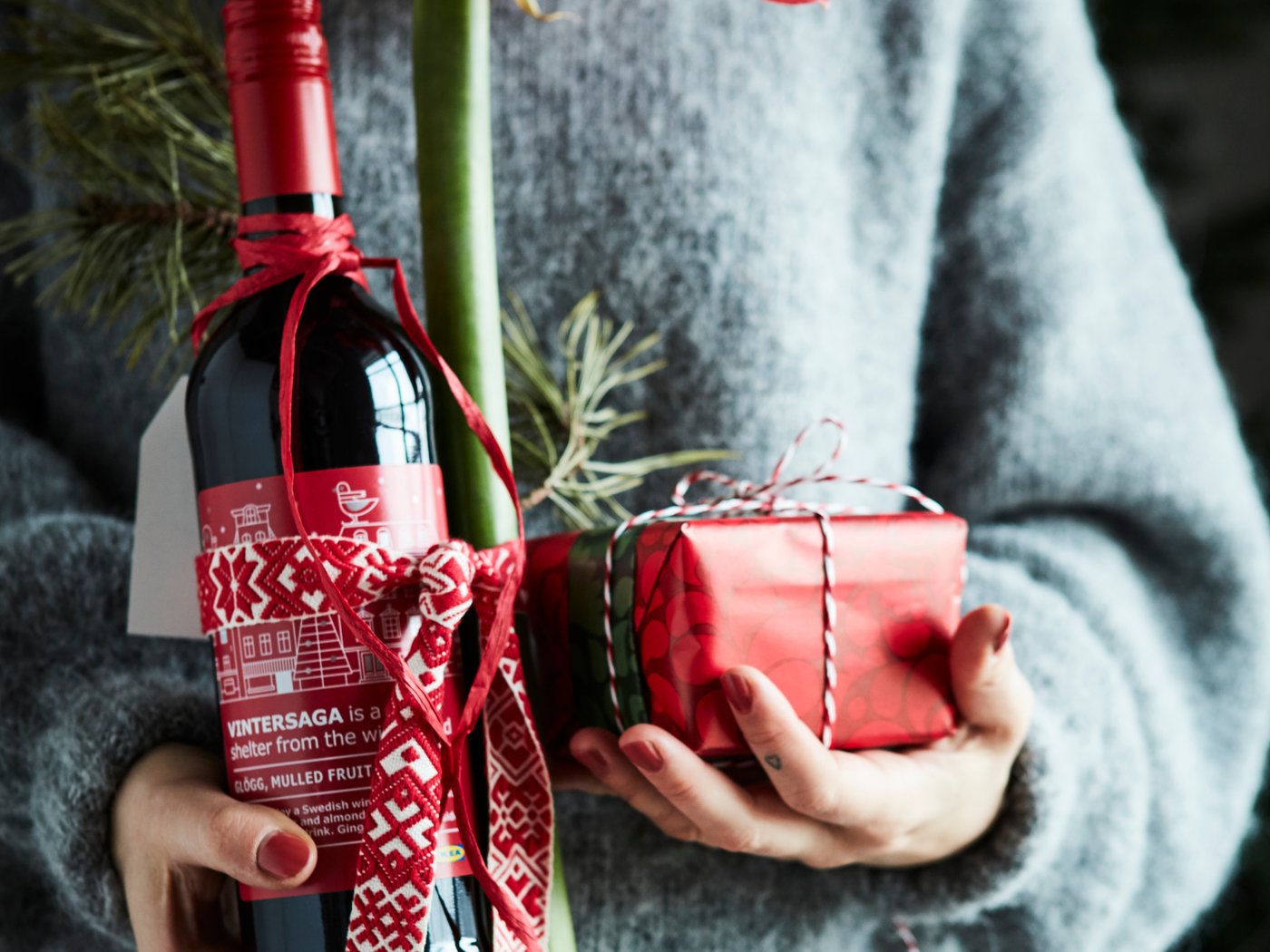 A person in a grey blue sweater holding a bottle of glögg and a small gift wrapped in a red wrapping paper.
