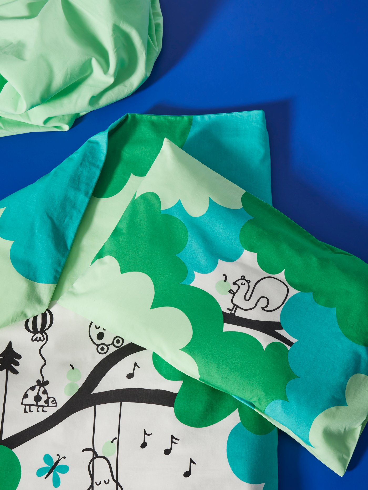 GRÖNFINK Bettwäsche mit buntem Design: Bäume mit grünen und blauen Blättern, ein Eichhörnchen, musikalische Noten und kleine Illustrationen auf blauem Hintergrund.