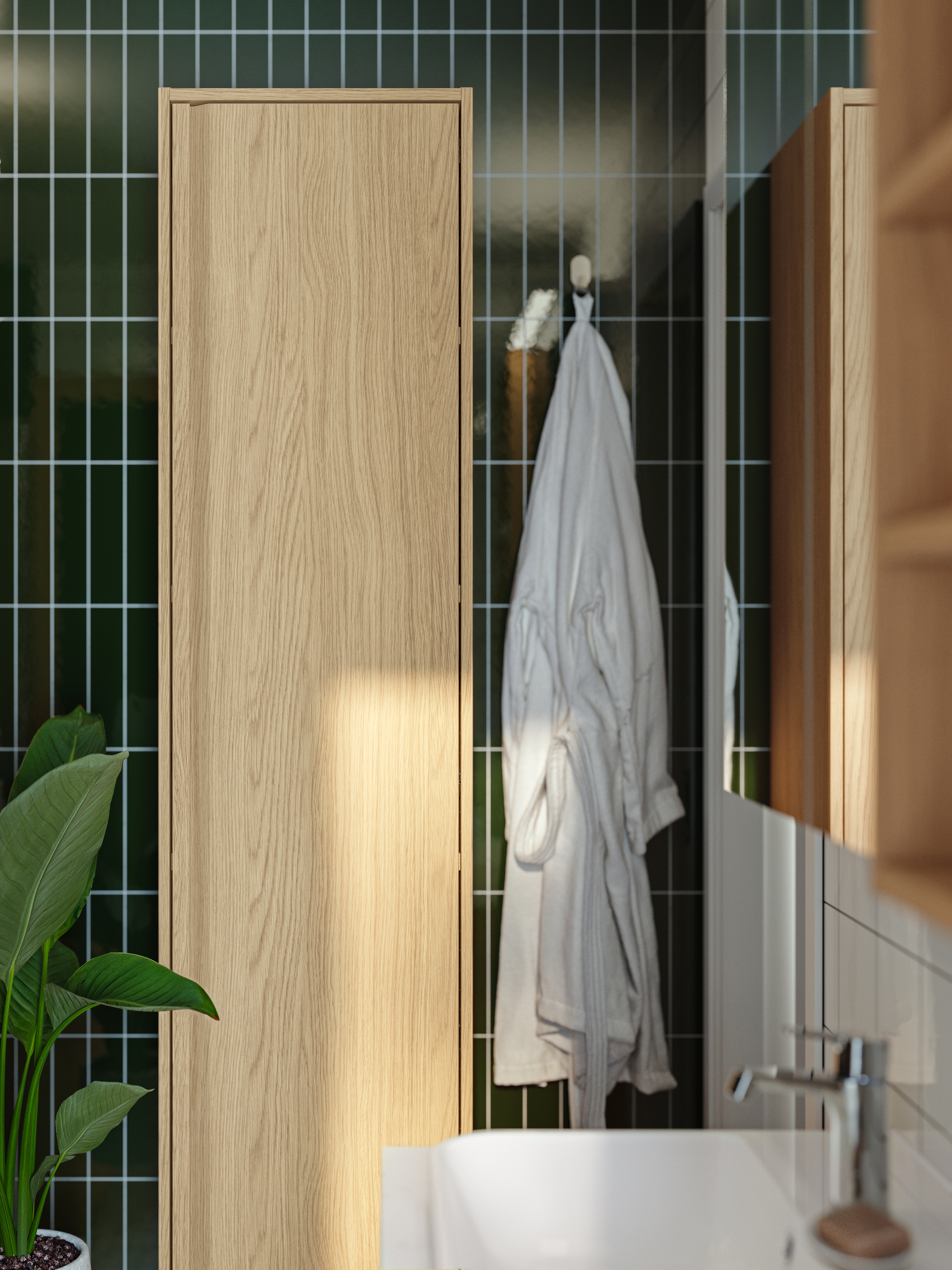An ÄNGSJÖN high cabinet with a door in oak effect is mounted on a wall with a white bathrobe handing on a hook next to it.
