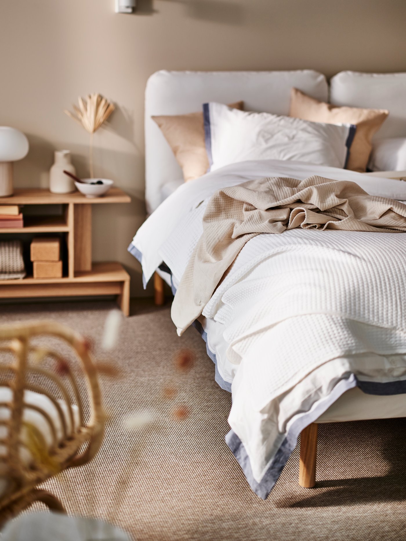 A white SAGESUND upholstered bed in a beige room with white/blue TÅTELSMYGARE bed linen and white VÅRELD bedspread on top.