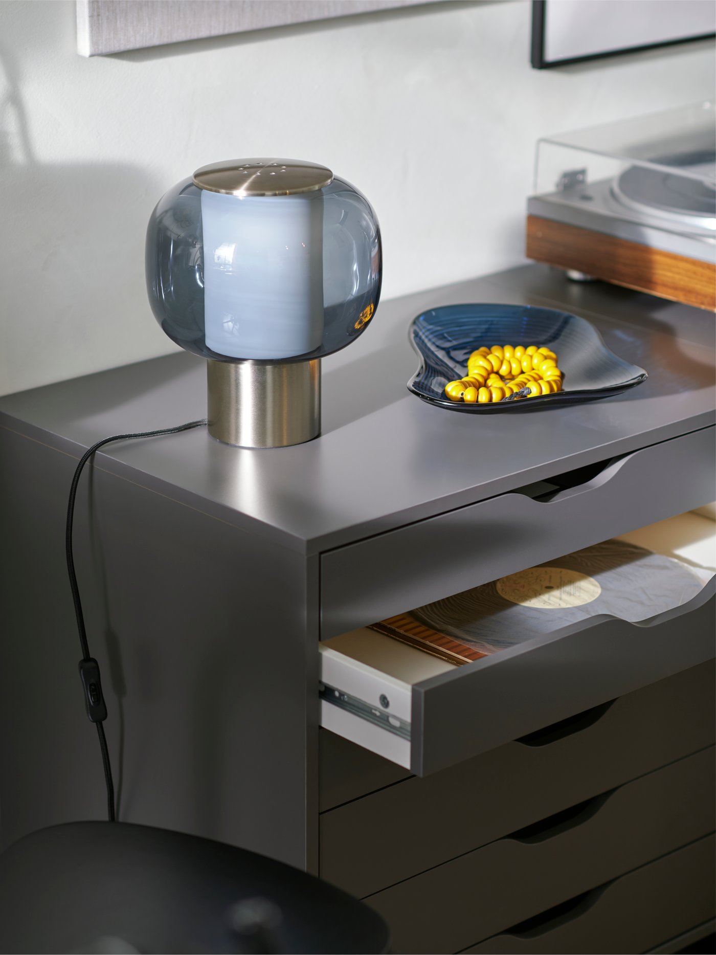 A HÖGVIND table lamp and a blue HÄCKPOPPEL decorative bowl with a yellow wooden necklace on a dark grey ALEX drawer unit.