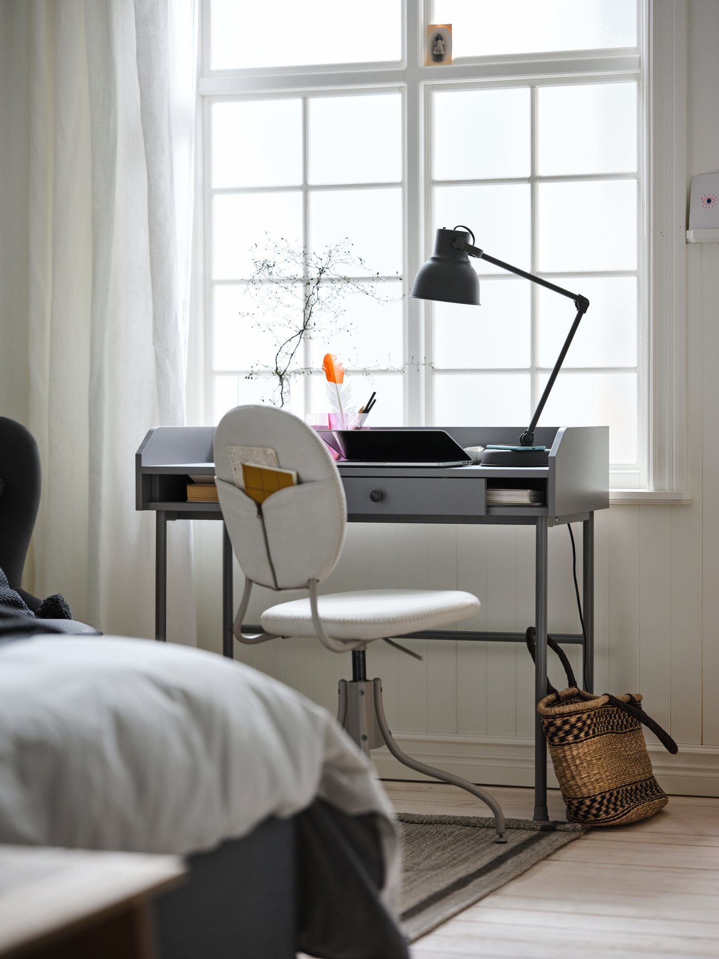 By a large, gridded window stands a beige BJÖRKBERGET chair and a grey HAUGA desk with a dark-grey HEKTAR work lamp on it.